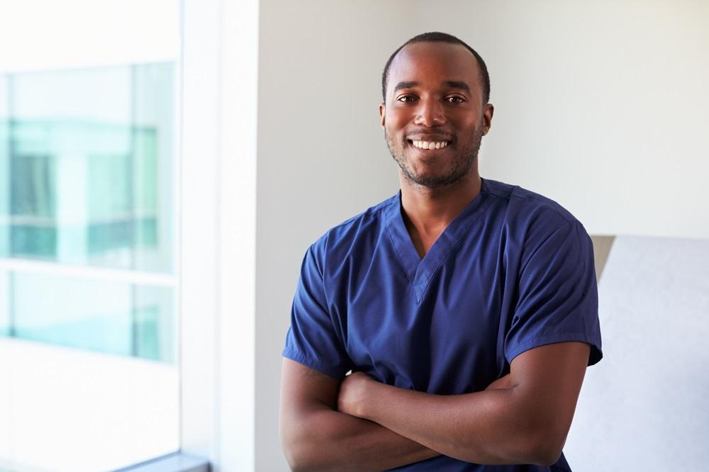 A young Black man dressed in purple scrubs smiling and standing with his arms folded.