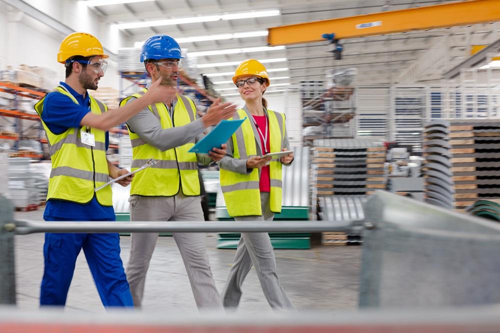 A group of workers walking through a factory with clipboards, hard hats, and reflective vests