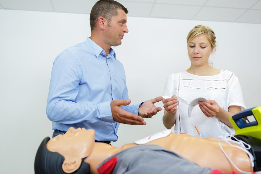 A white man in a blue button-up demonstrates how to use an AED to a white woman with an example mannequin in the foreground.